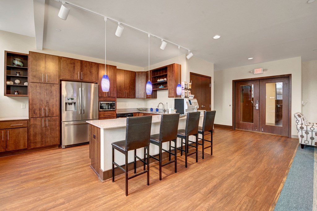 Avenir Apartments Community Kitchen with wooden cabinets, stainless steel appliances, and a central island with four bar stools. Blue pendant lights hang above, creating a warm, inviting ambiance.