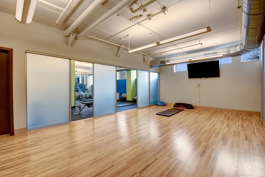 Spacious gym at Avenir Apartments with wooden floor, exercise mats, and yoga equipment. Frosted glass doors reveal gym machines. Bright lighting creates a clean, modern tone.