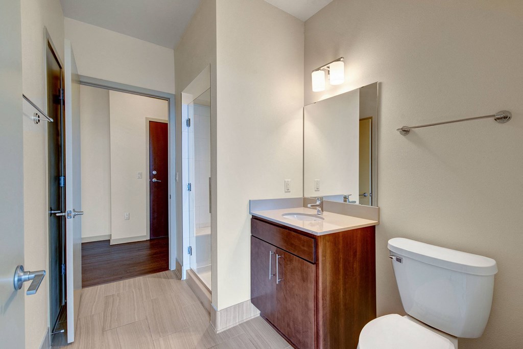 Modern bathroom with beige walls, wood vanity, mirror, and a toilet. Soft lighting from above creates a clean, calm atmosphere at Avenir Apartments.