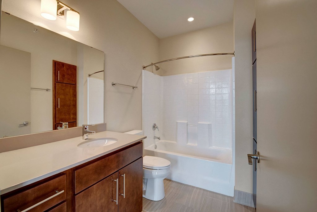 Modern bathroom at Avenir Apartments with beige walls, large mirror, wooden vanity, white countertop, and bathtub with tiled shower. Sleek, clean, and minimalist design.