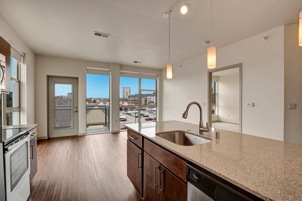 Modern apartment kitchen with granite countertops, dark wood cabinets, and stainless steel appliances. Bright light from large windows reveals a distant cityscape. Avenir Apartment Homes