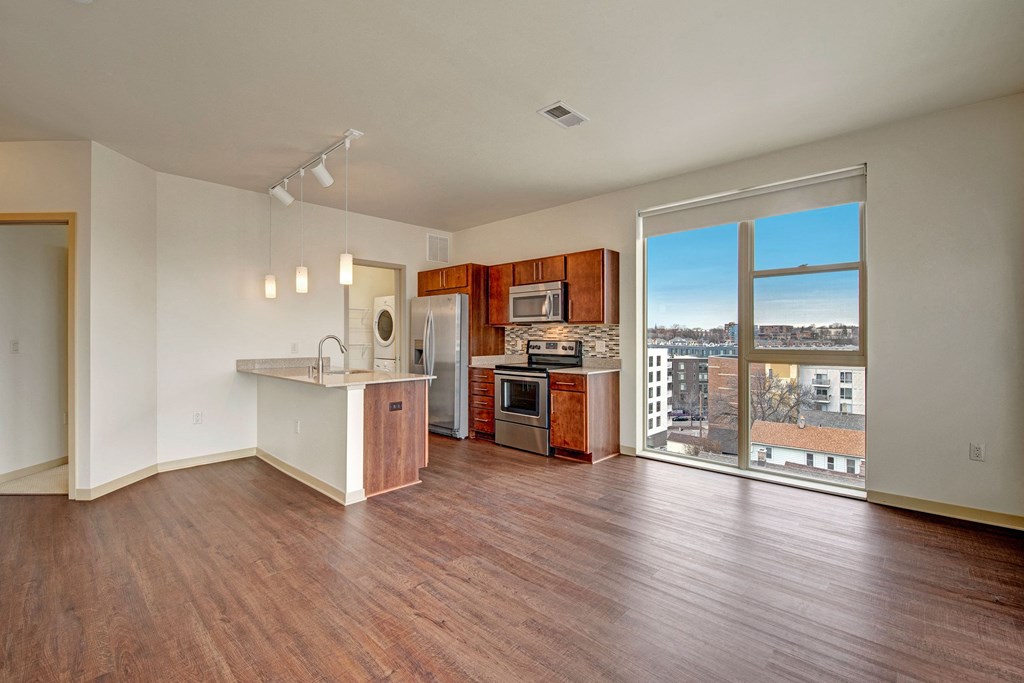 Modern apartment at Avenir Apartments with wooden floors, an open kitchen featuring stainless steel appliances, pendant lights, and a large window with city views.