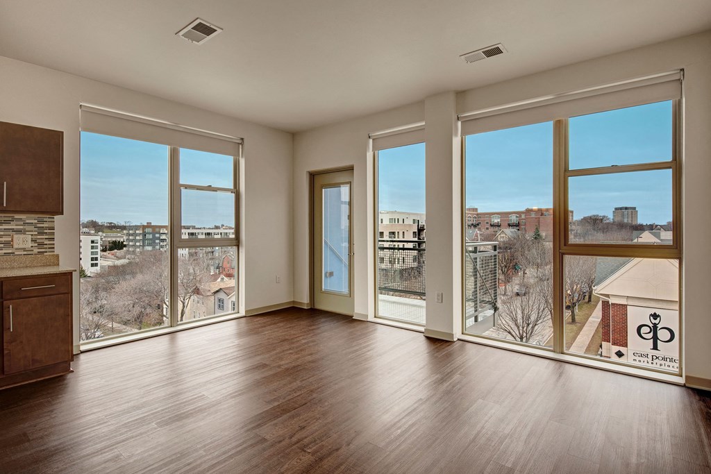 Bright, empty room with large windows showcasing an urban view. Hardwood floors, modern kitchen cabinets, and a calm ambiance at Avenir Apartments.