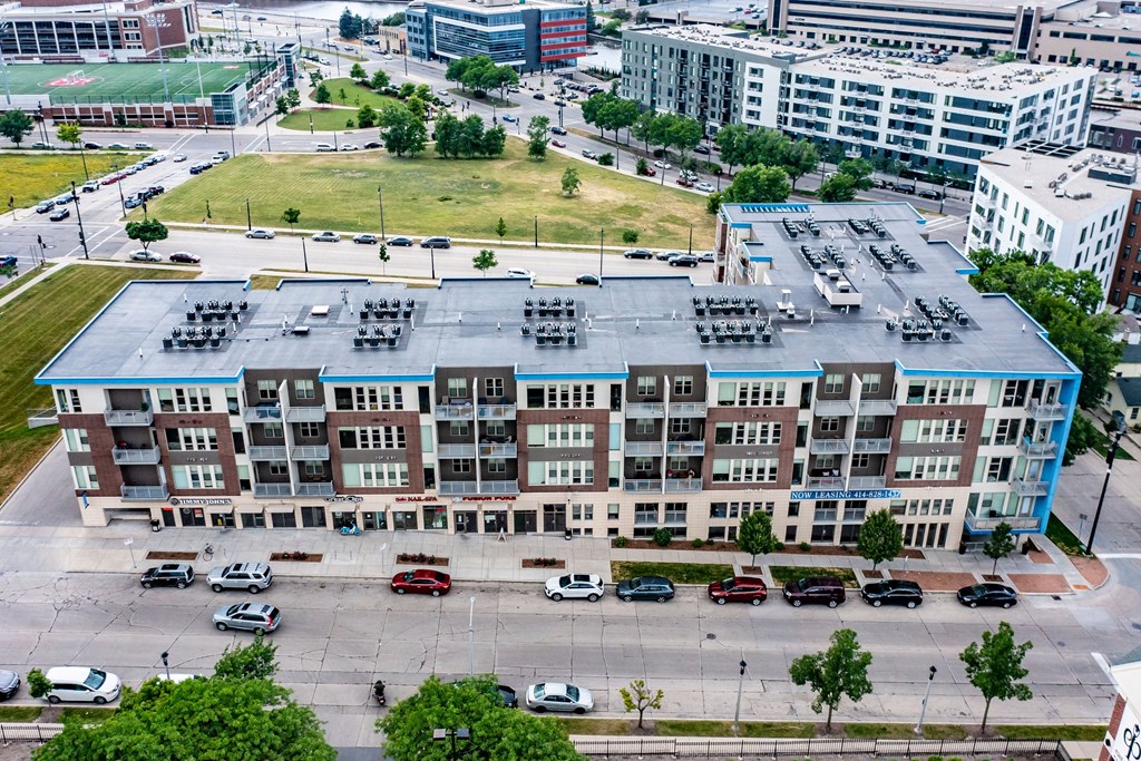 Aerial view of modern four-story Avenir apartments with a flat roof, surrounded by urban roads and patches of green space. The scene is lively and bustling.