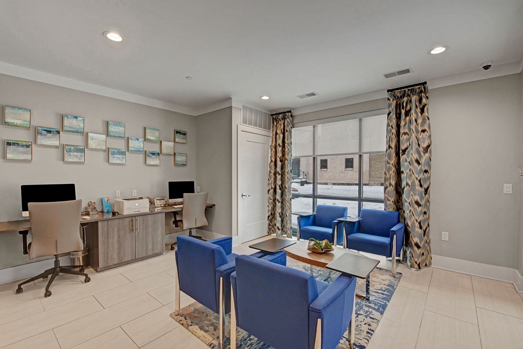 Modern business center with two workstations, computers, and printer on left. Blue chairs around tables in center, large window with patterned curtains behind at River House Apartments.