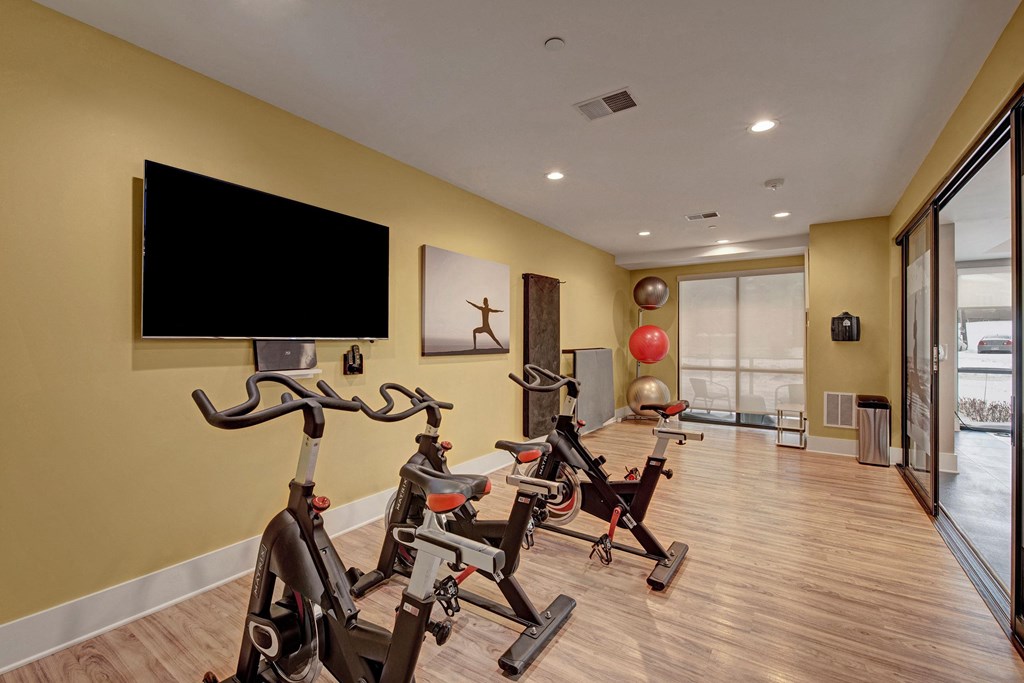 A modern home gym with three stationary bikes, a wall-mounted TV, a yoga poster, gym balls, and large windows, creating a focused, serene atmosphere at River House Apartments.