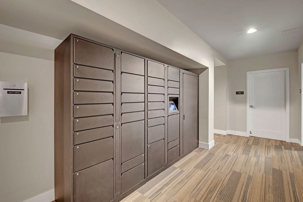 A hallway with a row of dark, numbered parcel lockers and a “Rent Drop” box on the left wall. Neutral tones and a closed white door at River House Apartments.