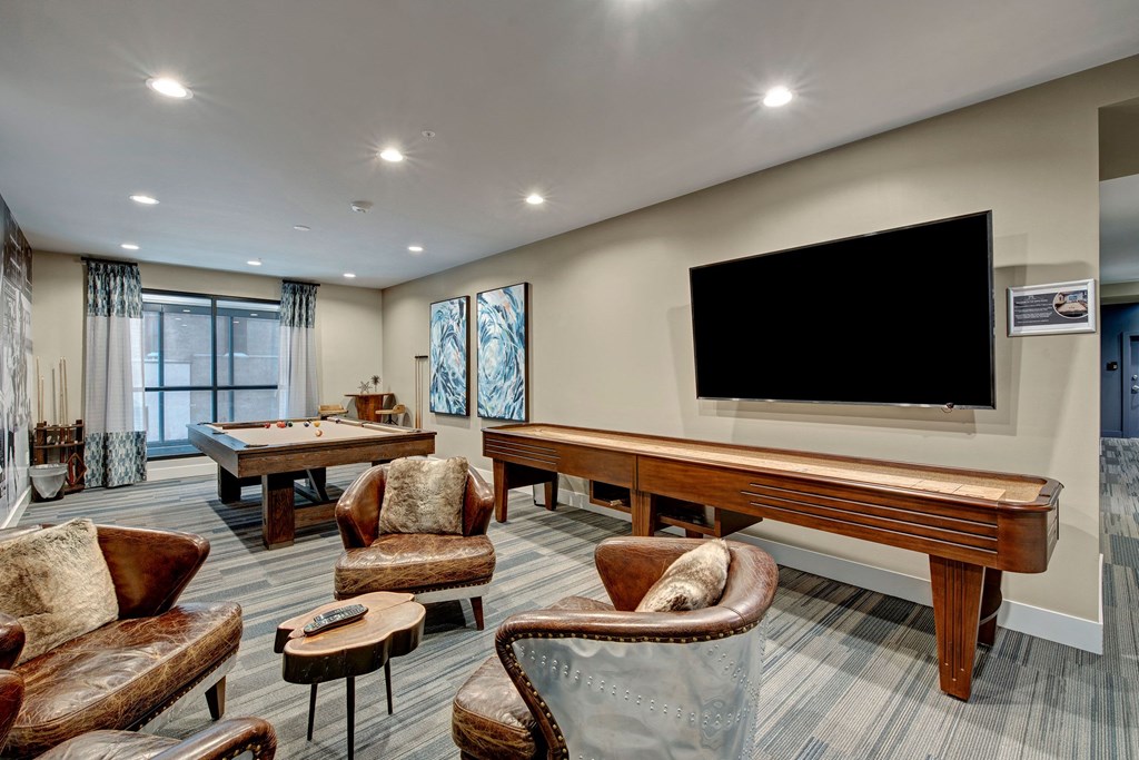 Cozy community room with a pool table, shuffleboard, leather chairs, and a large TV at River House Apartments.