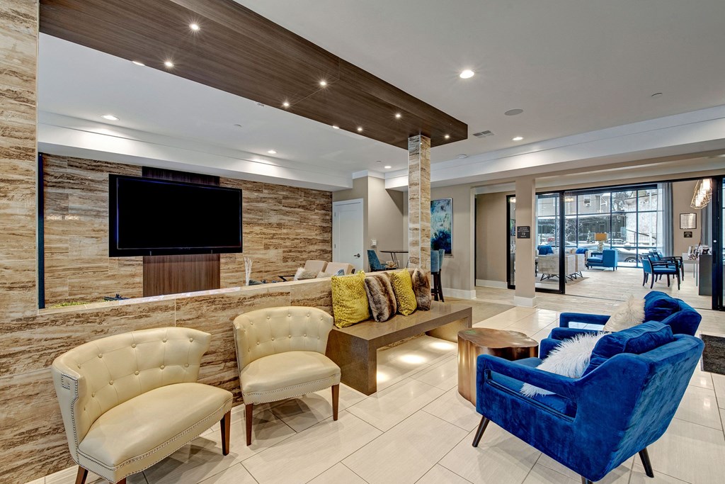Modern lobby with a sleek, contemporary design. Features cream chairs, a plush blue sofa, colorful cushions, wood accents, and a large TV at River House Apartments.