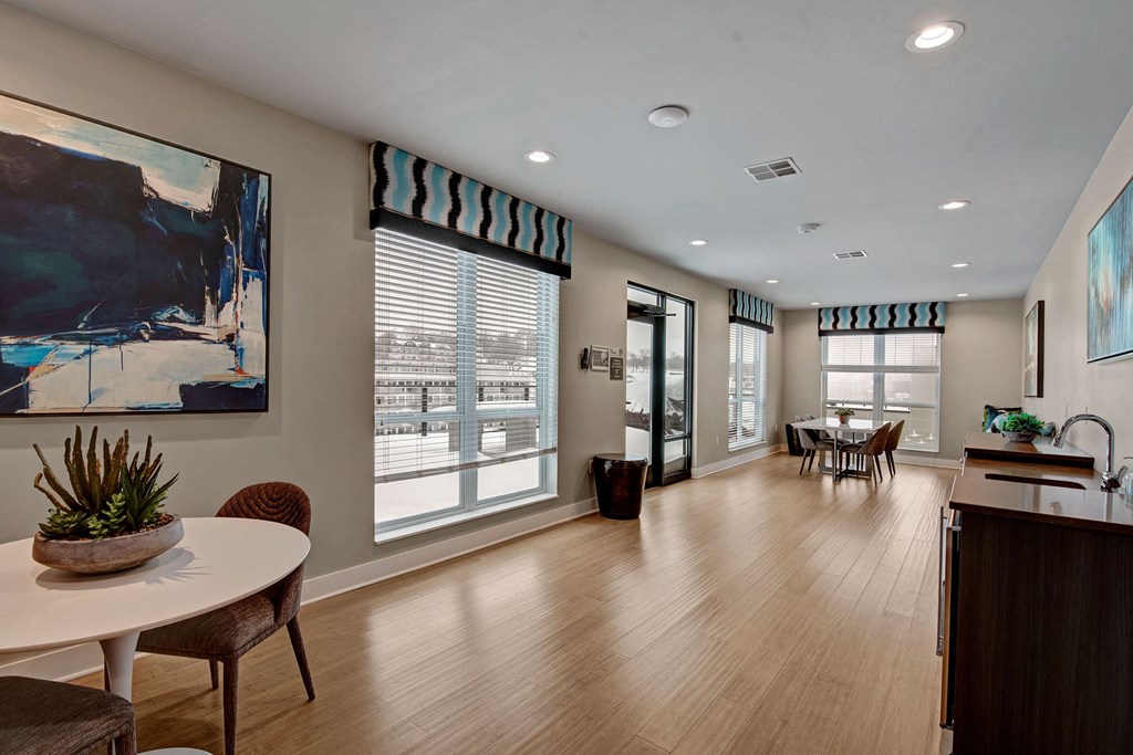 Modern community room with wood flooring and large windows covered by patterned valances. Features abstract art, a round table, chairs, and plants at River House Apartments.
