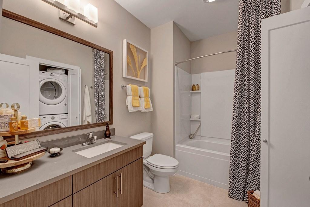 Modern bathroom with wood vanity, large mirror, and stacked washer-dryer. Bathtub with patterned curtain, towels, and framed art create a cozy feel at River House Apartments.