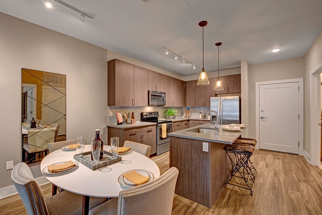 Modern kitchen with sleek wood cabinets, stainless steel appliances, island with barstools, and a round dining table set for four. Warm lighting and neutral tones create a cozy atmosphere at River House Apartments.
