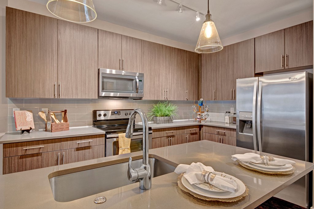 Modern kitchen with wood cabinets, stainless steel appliances, and a double-door fridge. Central island has a sink and two place settings with napkins. Warm lighting creates an inviting atmosphere at River House Apartments.