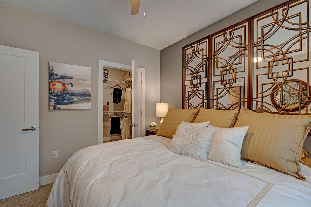 Elegant bedroom with a large, pillowed bed, gold accents, and artistic wall panels. Open closet visible, adding a warm, inviting feel at River House Apartments.