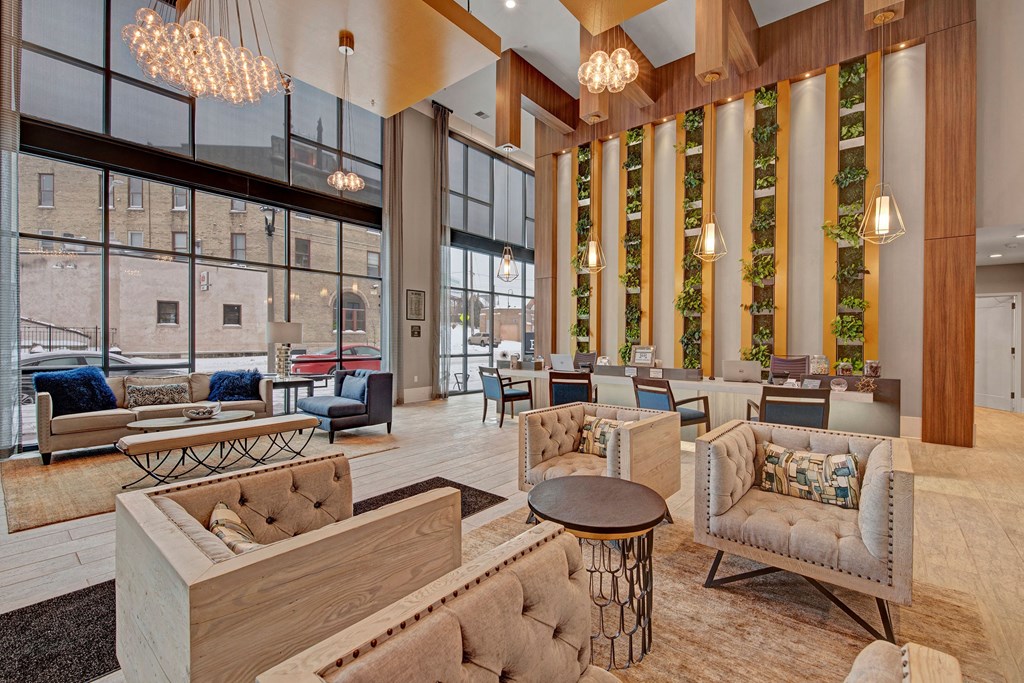 Modern apartment lobby featuring cozy seating with tufted armchairs and sofas, large windows, hanging chandeliers, greenery wall, and a calm ambiance at River House Apartments.