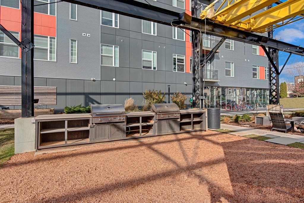 Outdoor area with two metal grills under a modern structure, adjacent to a gray building. Bicycles and lounge chairs are visible. Bright, sunny day at Stitchweld Apartments.