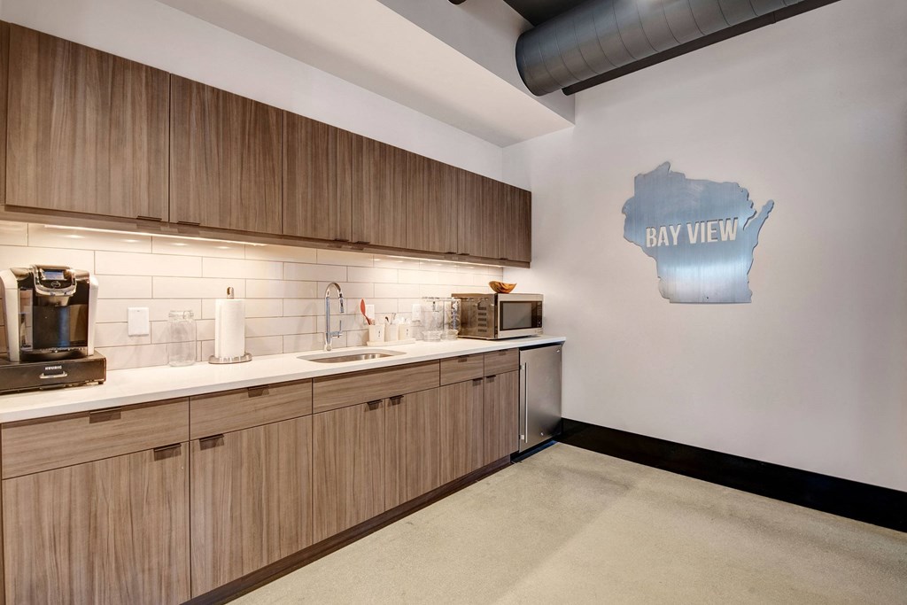 Modern kitchen with wooden cabinets, white countertops, and a stainless steel sink. A sign shaped like Wisconsin says "eat well" on the wall, creating a welcoming vibe at Stitchweld Apartments.