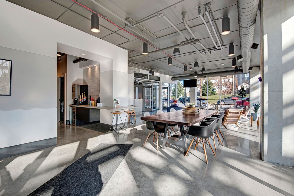 Modern cafe interior with large windows and natural light. Features a long wooden table with black chairs, a coffee station, and minimalist decor at Stitchweld Apartments.