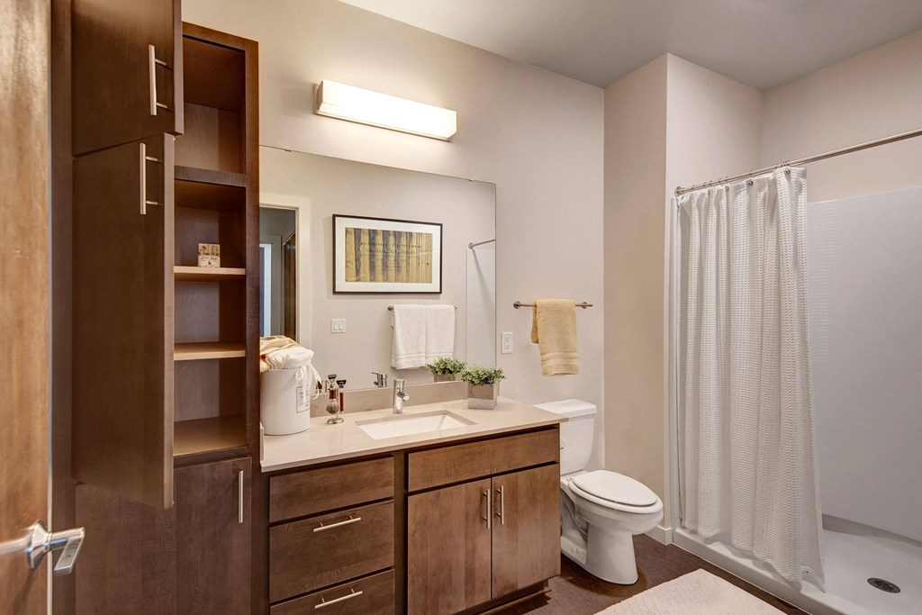 Modern bathroom with wooden cabinets, white countertop, and sink beside a toilet. A shower with a white curtain is on the right at Stitchweld Apartments.