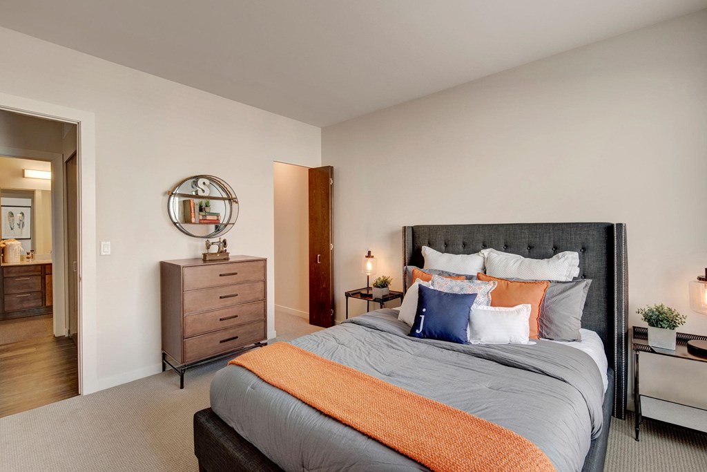 Modern bedroom with a plush gray bed, layered pillows, and an orange throw blanket. A wooden dresser and round mirror are on the left at Stitchweld Apartments.