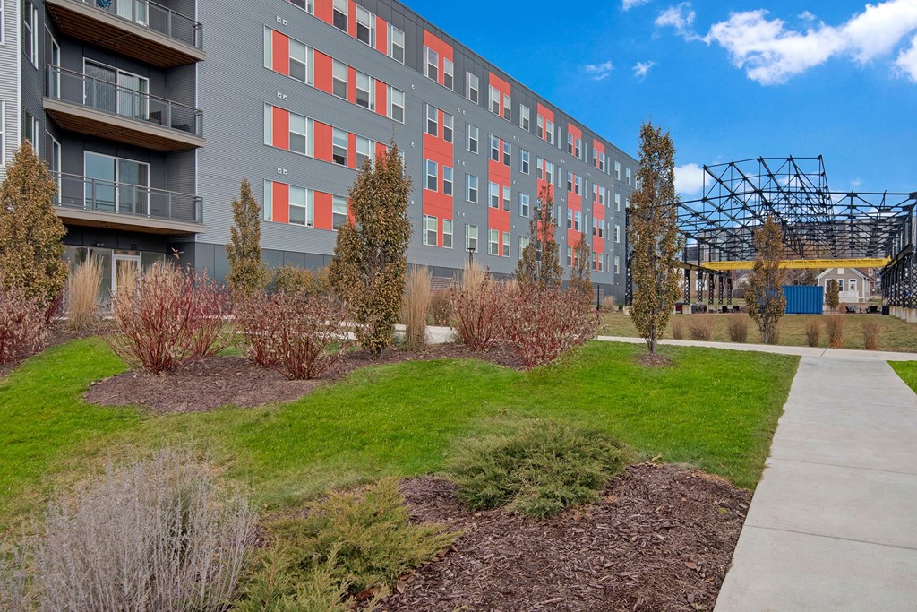 Modern apartment building with gray and red panels, surrounded by landscaped greenery. A pathway and metal structure are visible under a clear blue sky at Stitchweld Apartments.