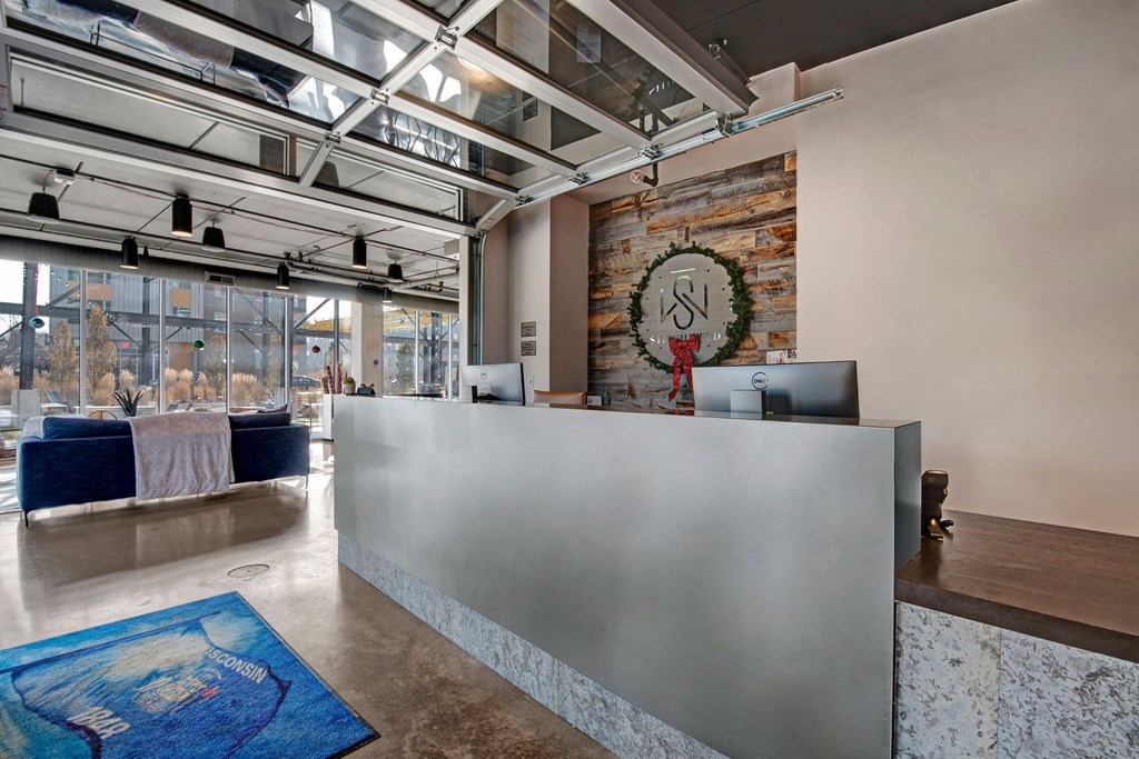 Modern reception desk with minimalist design, featuring two computers. A logo with a holiday wreath is on the wall. Large windows and blue couches add a welcoming ambiance at Stitchweld Apartments.