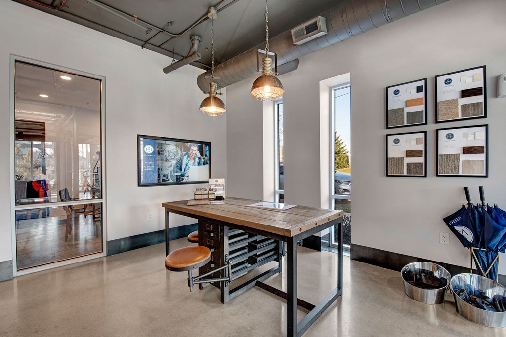 Modern office lobby with industrial decor. A wooden table and stool set is centered under two pendant lights. Sample boards and umbrellas line the walls, creating a welcoming atmosphere at Stitchweld Apartments.