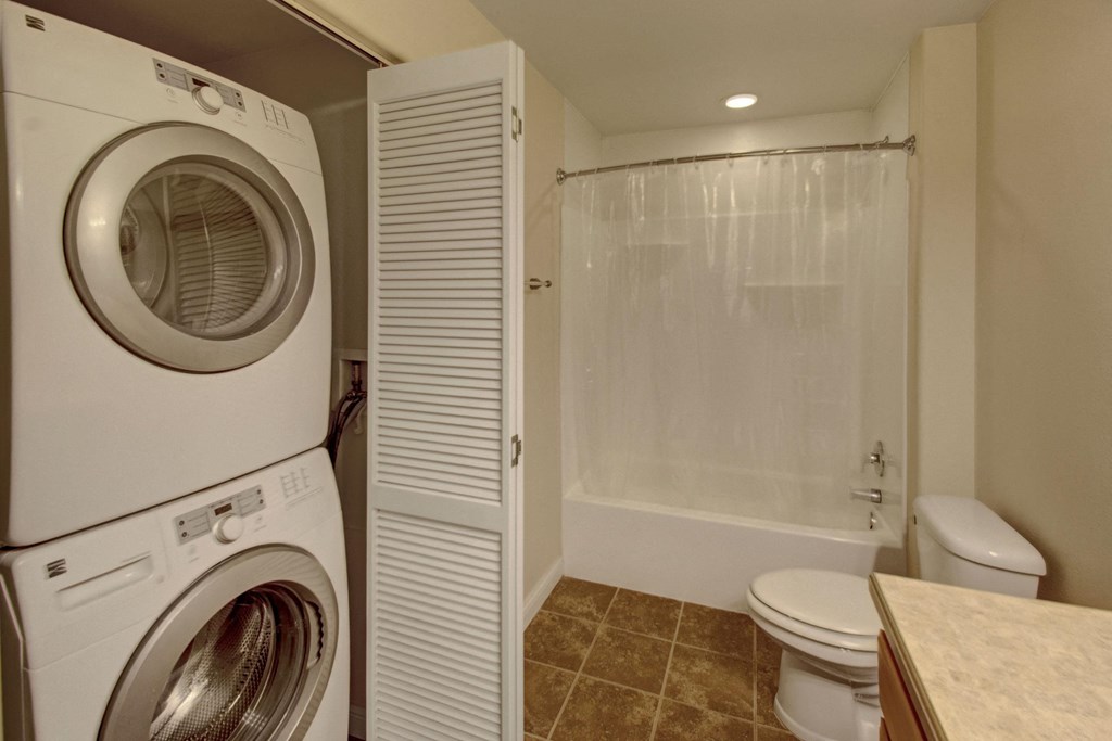 Compact laundry and bathroom space here at Confluence at the Harvest Hills Apartment Homes with stacked washer and dryer on the left, white bathtub with a curtain, toilet, and tiled floor. Clean and functional.