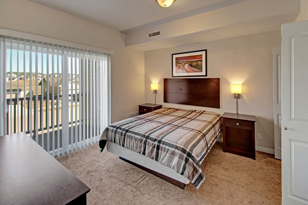 A cozy bedroom here at Confluence at the Harvest Hills Apartment Homes with a plaid-patterned bed, flanked by two nightstands and wall lamps. Large window with vertical blinds offers natural light.