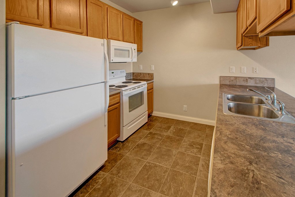 Compact kitchen here at Confluence at the Harvest Hills Apartment Homes with wooden cabinets, white appliances including fridge, stove, and microwave. Spacious brown tiled countertop with double sink. Neutral tones convey simplicity.