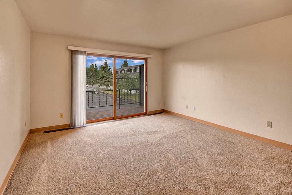 An empty living room with beige carpet and white walls. A large sliding glass door opens to a balcony with a view of trees and buildings, conveying a serene atmosphere. The Alaskan Apartments.