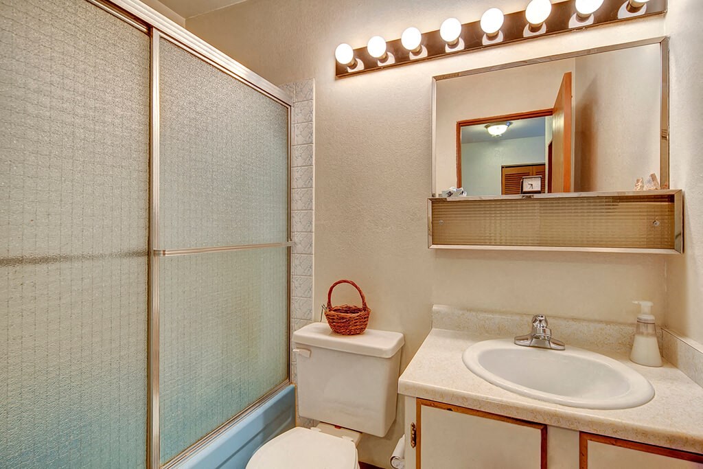 Compact bathroom with a glass shower door, white toilet, and beige countertop with a sink. A wicker basket is on the toilet tank and bright lights above a mirror. The Alaskan Apartments.