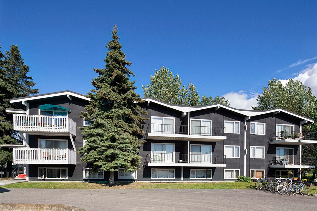 Three-story apartment building with dark siding, white balconies, and a teal umbrella. Surrounded by evergreens and blue sky. Bicycles are parked nearby. The Alaskan Apartments.