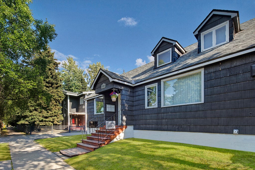 A quaint black house with white trim under a clear blue sky, featuring dormer windows and a flower pot. The sign reads “Leasing Office,” evoking a welcoming vibe. The Alaskan Apartments.