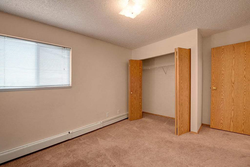Empty bedroom here at Aspen Terrace Apartment Homes with beige walls and carpet. A window with blinds on the left, open closet with wooden doors in the center, and a closed door on the right.