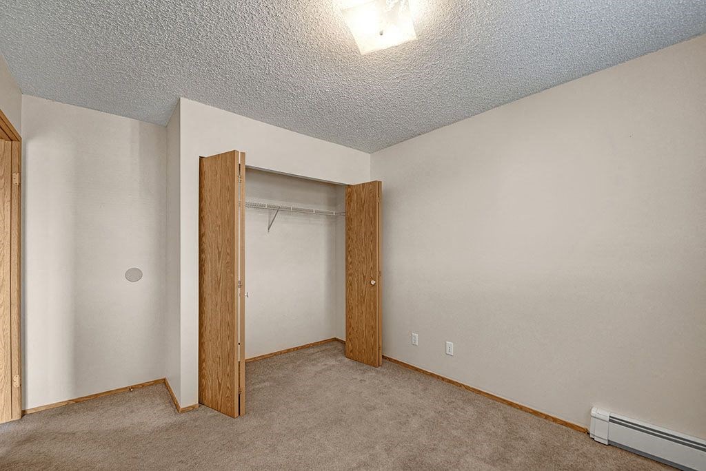Empty beige bedroom here at Aspen Terrace Apartment Homes with open wooden closet doors revealing a shelf and rod. The room has a carpeted floor, textured ceiling, and a bright light fixture.