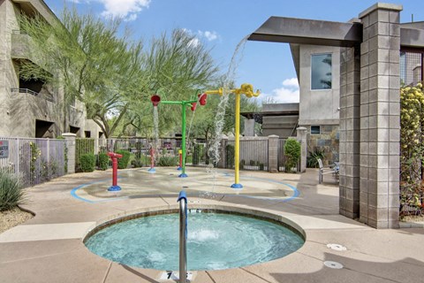 A vibrant children's splash pad here at Aspire at Pinnacle Peak Apartments with colorful water features and a small pool in a sunny outdoor setting. Surrounded by trees and concrete buildings.