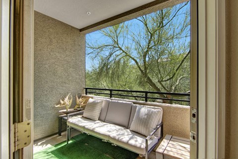 Cozy balcony with a cushioned sofa and striped pillows, overlooking a lush tree here at Aspire at Pinnacle Peak Apartments. Sunlight enhances the relaxed, natural ambiance.