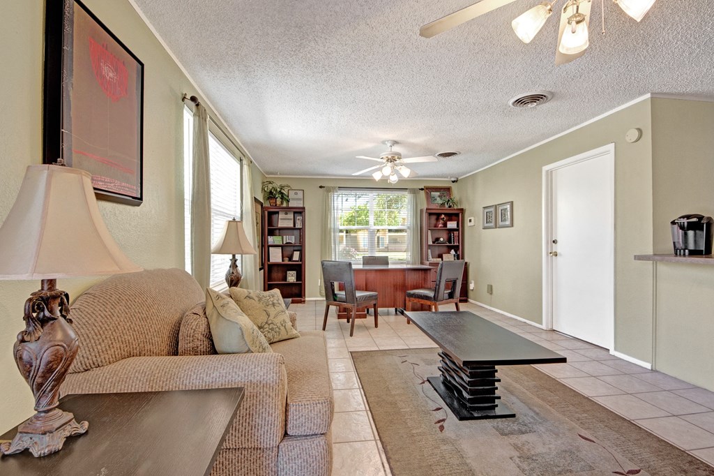 Cozy living room here at Avalon Springs Apartment Homes with beige walls, featuring a plush sofa, ornate lamps, and a sleek coffee table. A window-lit desk area with bookshelves adds warmth.