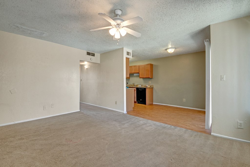 A spacious, empty living room here at Avalon Springs Apartment Homes with beige carpet and walls, featuring a ceiling fan with lights. The adjoining kitchen has wooden cabinets and wood flooring.