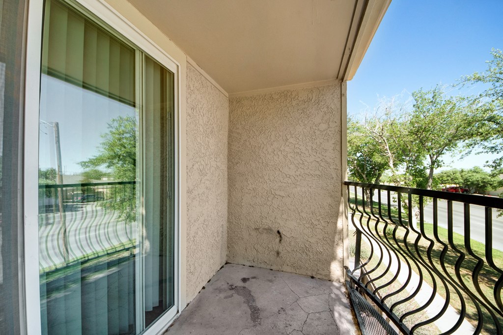 Small concrete balcony here at Avalon Springs Apartment Homes with textured beige walls and a sliding glass door on one side. Black metal railing overlooks a street lined with green trees.