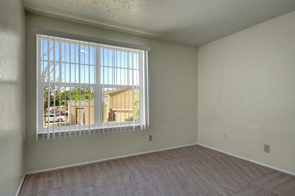 Empty bedroom here at Avalon Springs Apartment Homes with beige carpet and white walls, featuring a large window with vertical blinds on the left. Outside, greenery and buildings are visible. Bright and airy atmosphere.