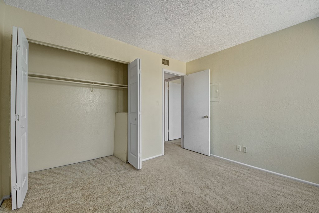 Empty room here at Avalon Springs Apartment Homes with beige carpet and light beige walls. Open closet doors reveal a hanging rod. An open door leads to another room, creating a neutral, calm space.