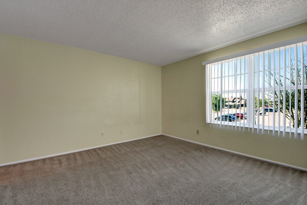 Empty bedroom here at Avalon Springs Apartment Homes with beige walls and carpet, large window with vertical blinds, and a view of parked cars and trees outside. Bright and minimalistic.