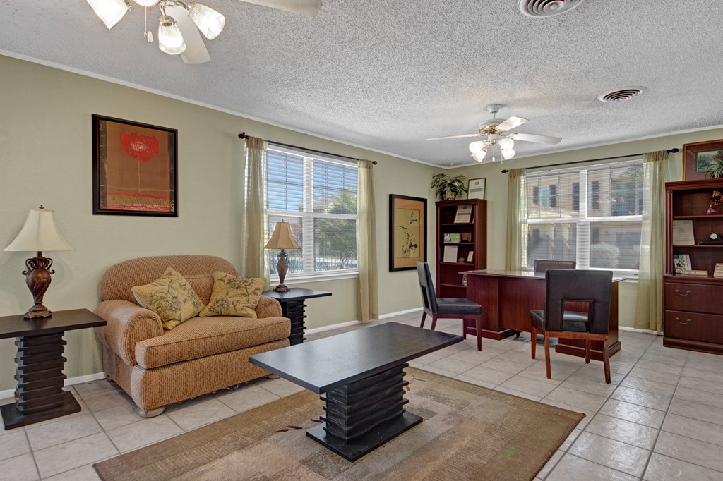 A cozy office room here at Avalon Springs Apartment Homes with a brown armchair, two decorative side tables, and a central dark wood coffee table. A desk with two chairs, bookcases, and large windows letting in ample light create an inviting atmosphere.