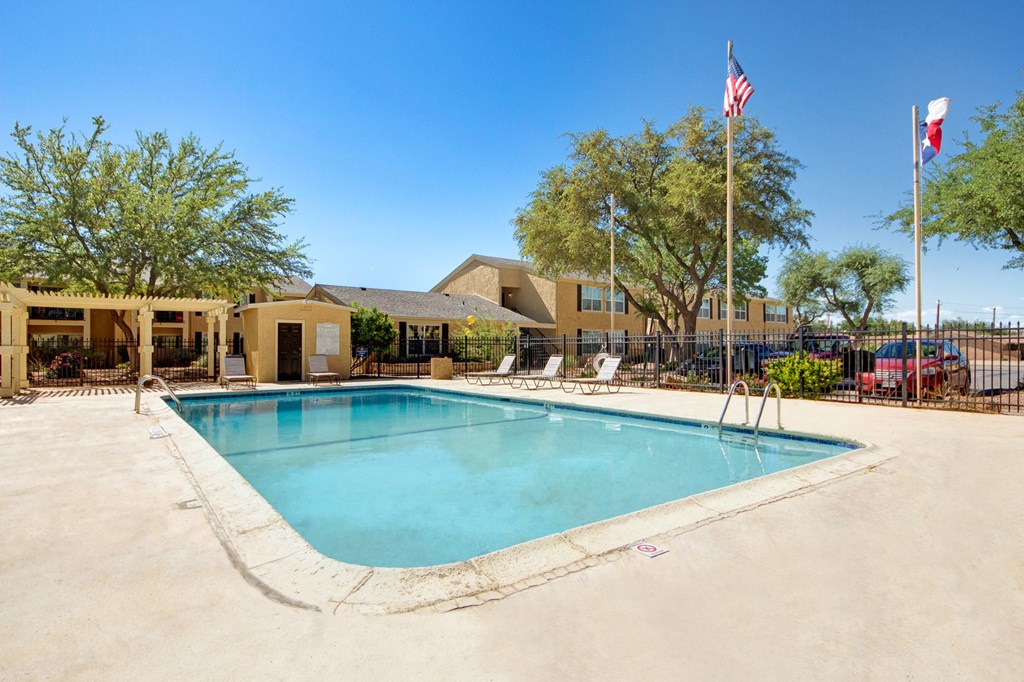 Outdoor pool area here at Avalon Springs Apartment Homes on a sunny day, surrounded by lounge chairs and trees. Flags on poles in the background, with a clear blue sky and adjacent building.