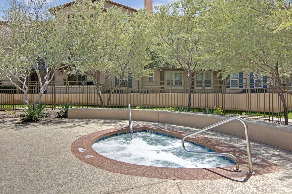 Outdoor hot tub here at Bela Rosa Apartments with bubbling water, surrounded by a textured concrete surface. Tall trees and a beige building with windows are in the background. Relaxed ambiance.