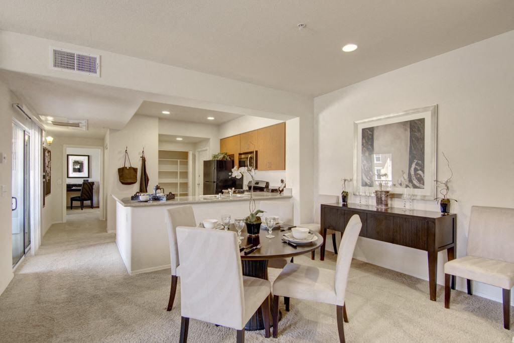 Bright, modern dining area here at Bela Rosa Apartments with a round table set for four, beige chairs, and elegant decor. Nearby, an open kitchen with wood cabinets and white walls.