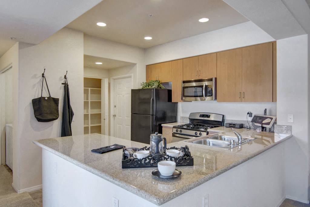 Modern kitchen here at Bela Rosa Apartments with beige granite countertops, stainless steel appliances, light wood cabinets, and neatly arranged utensils. Cozy and inviting atmosphere.