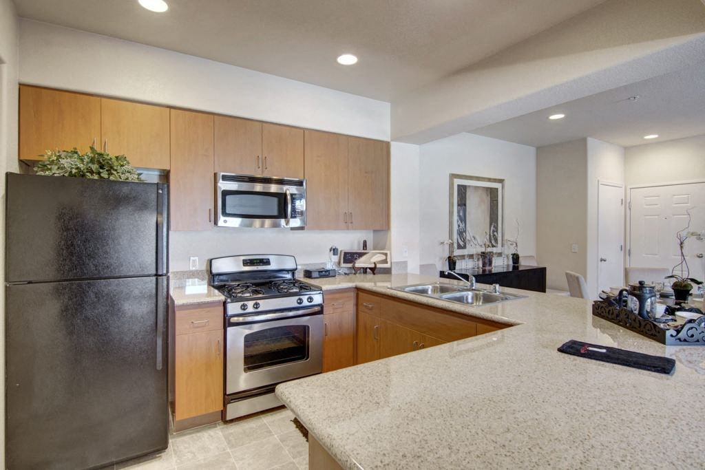 Modern kitchen here at Bela Rosa Apartments with wooden cabinets, granite countertops, stainless steel stove, and refrigerator. Bright lighting creates a clean, welcoming atmosphere.